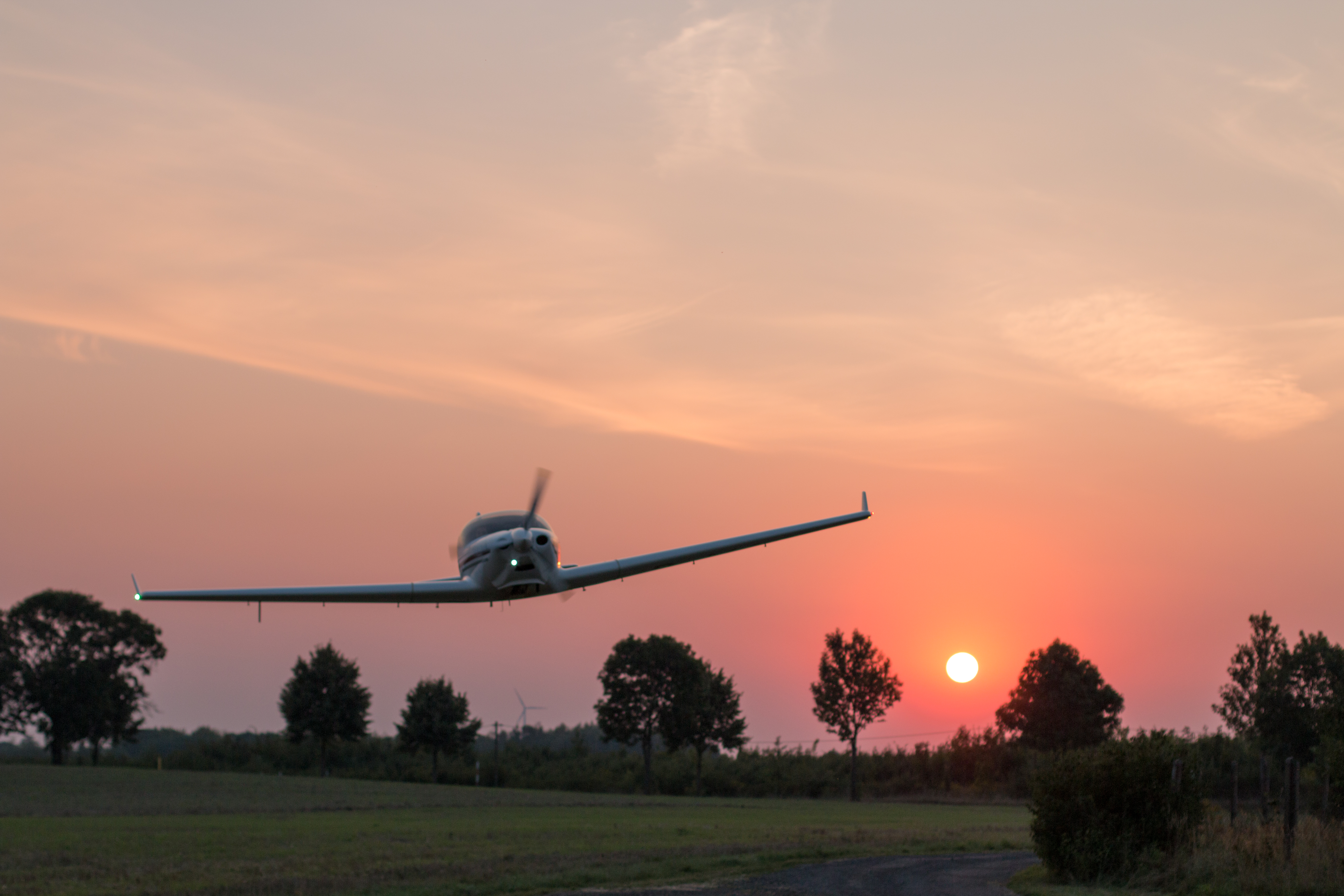 Ein Flugzeug fliegt tief über eine ländliche Gegend, während die Sonne am Horizont untergeht.