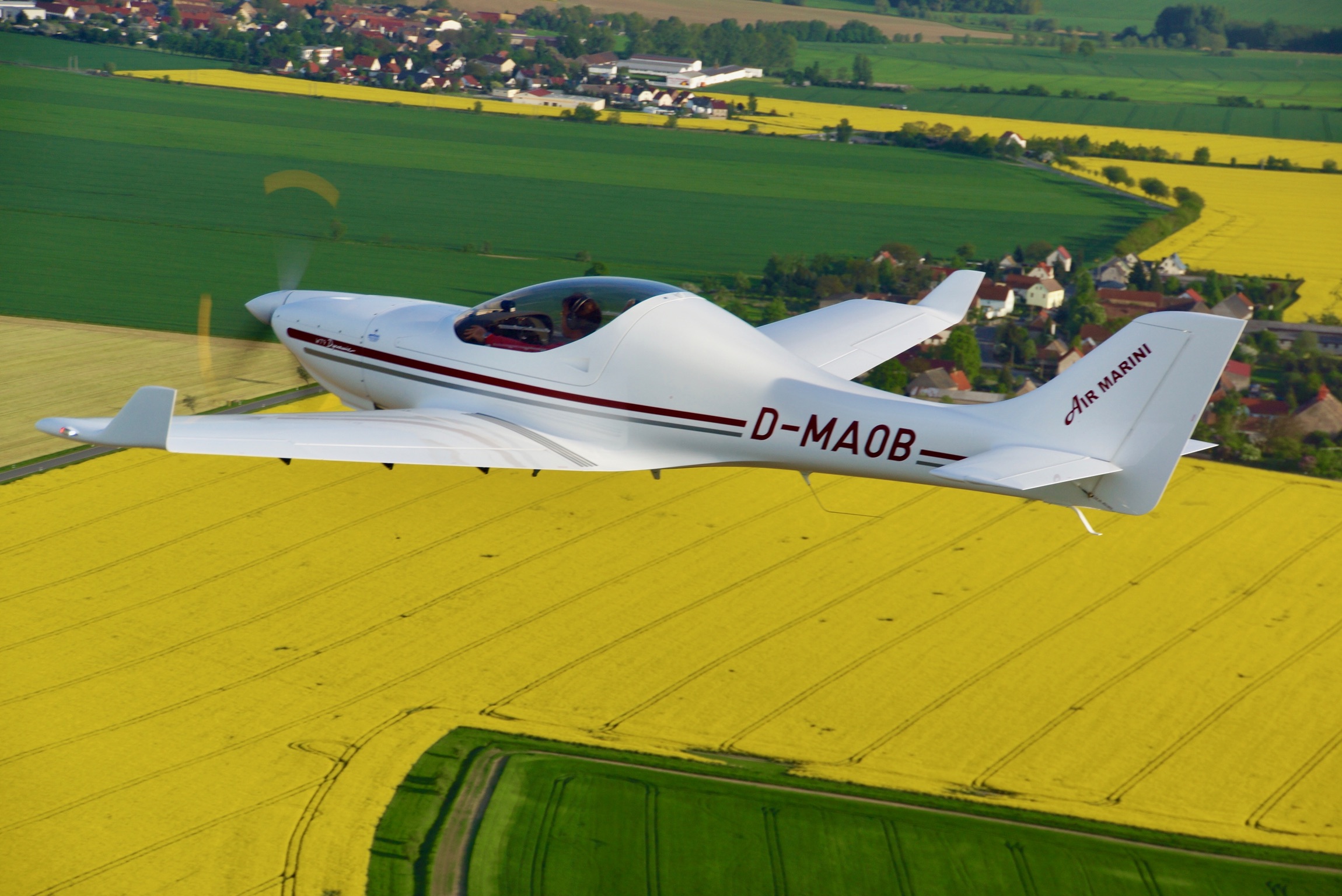 Ein weißes Leichtflugzeug mit der Kennzeichnung D-MA0B fliegt über gelbe und grüne Felder mit einem Dorf im Hintergrund