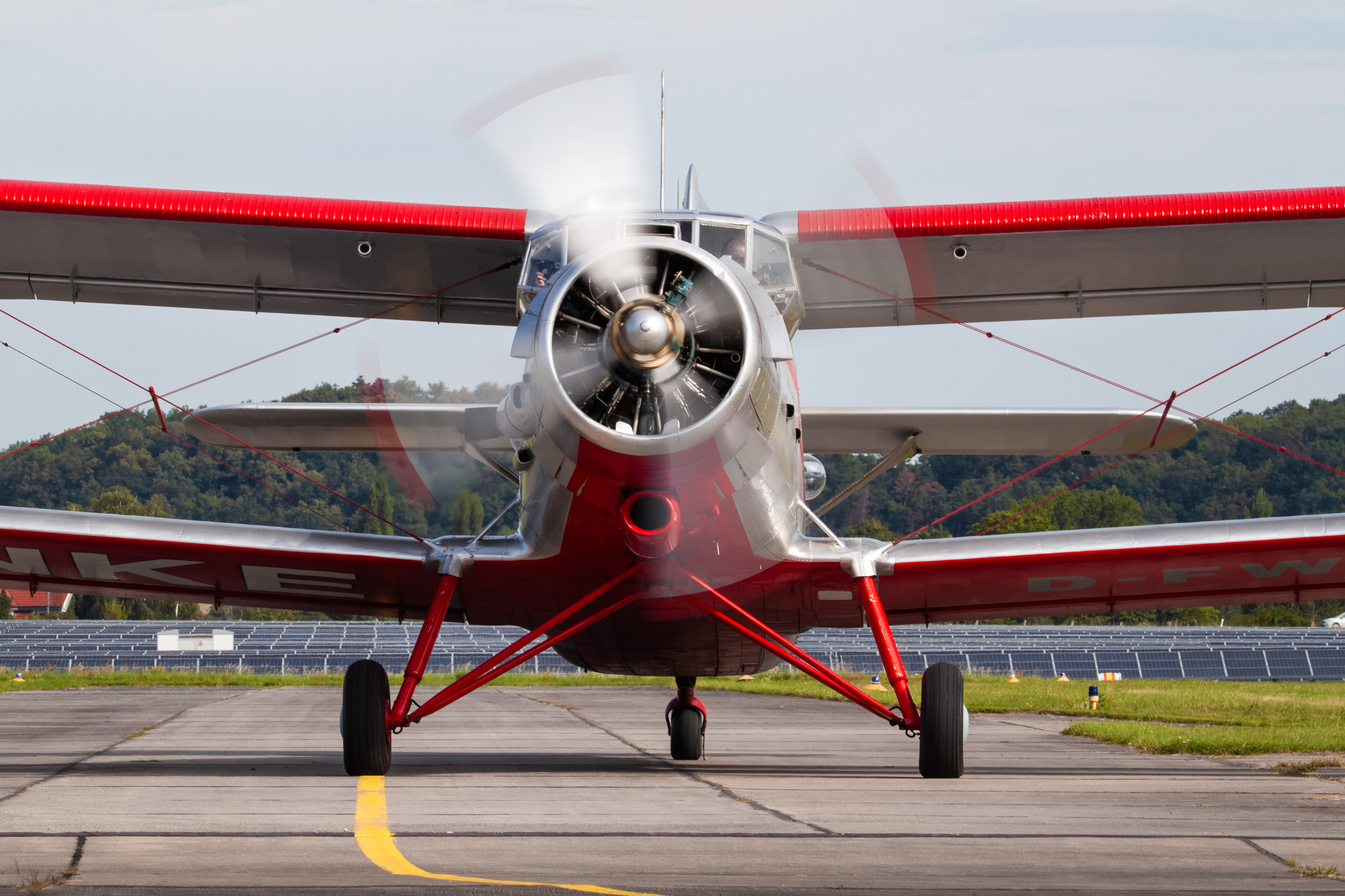 Vorderansicht eines rot-weißen Propellerflugzeugs auf der Landebahn mit drehendem Propeller vor einem bewaldeten Hintergrund