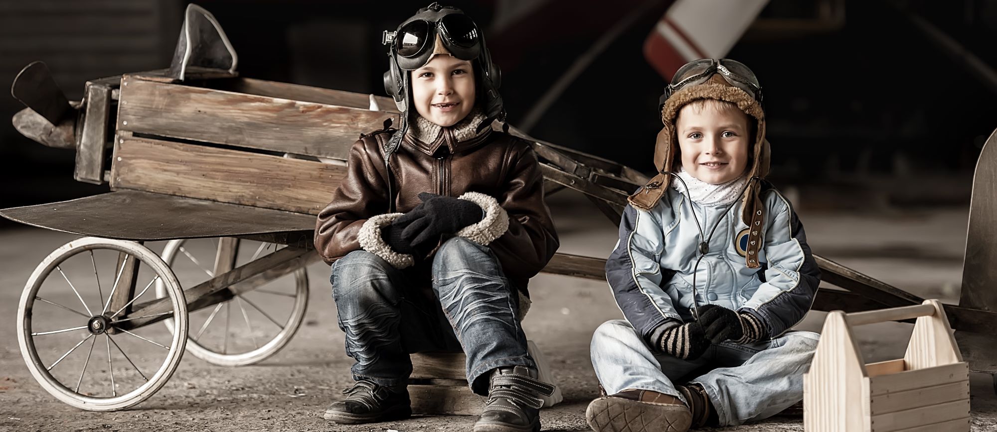 Zwei Kinder mit Fliegerhauben und Schutzbrillen vor einem hölzernen Flugzeug