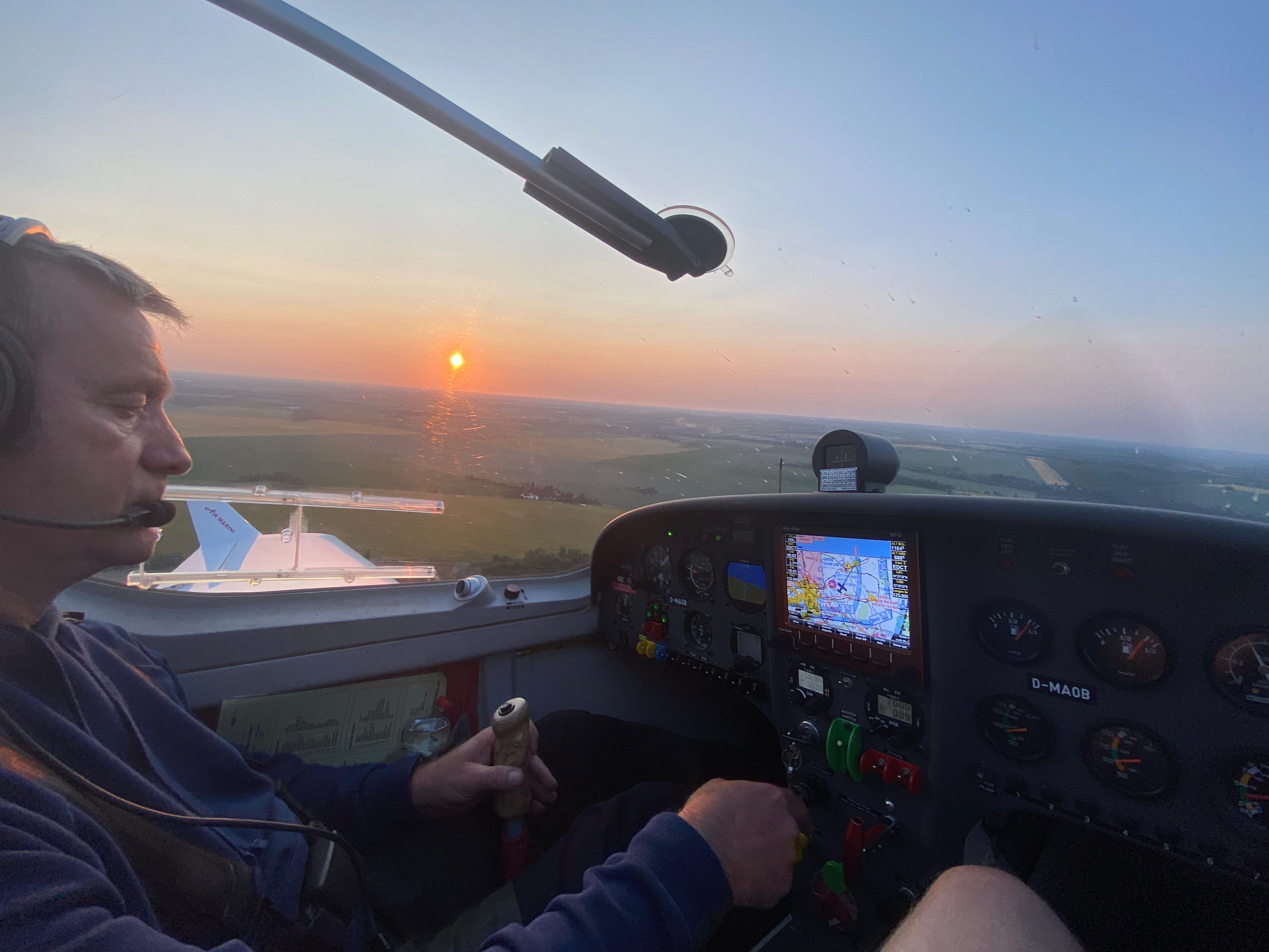 Ein Pilot steuert ein kleines Flugzeug am Abendhimmel, der Sonnenuntergang ist durch das Cockpitfenster sichtbar.