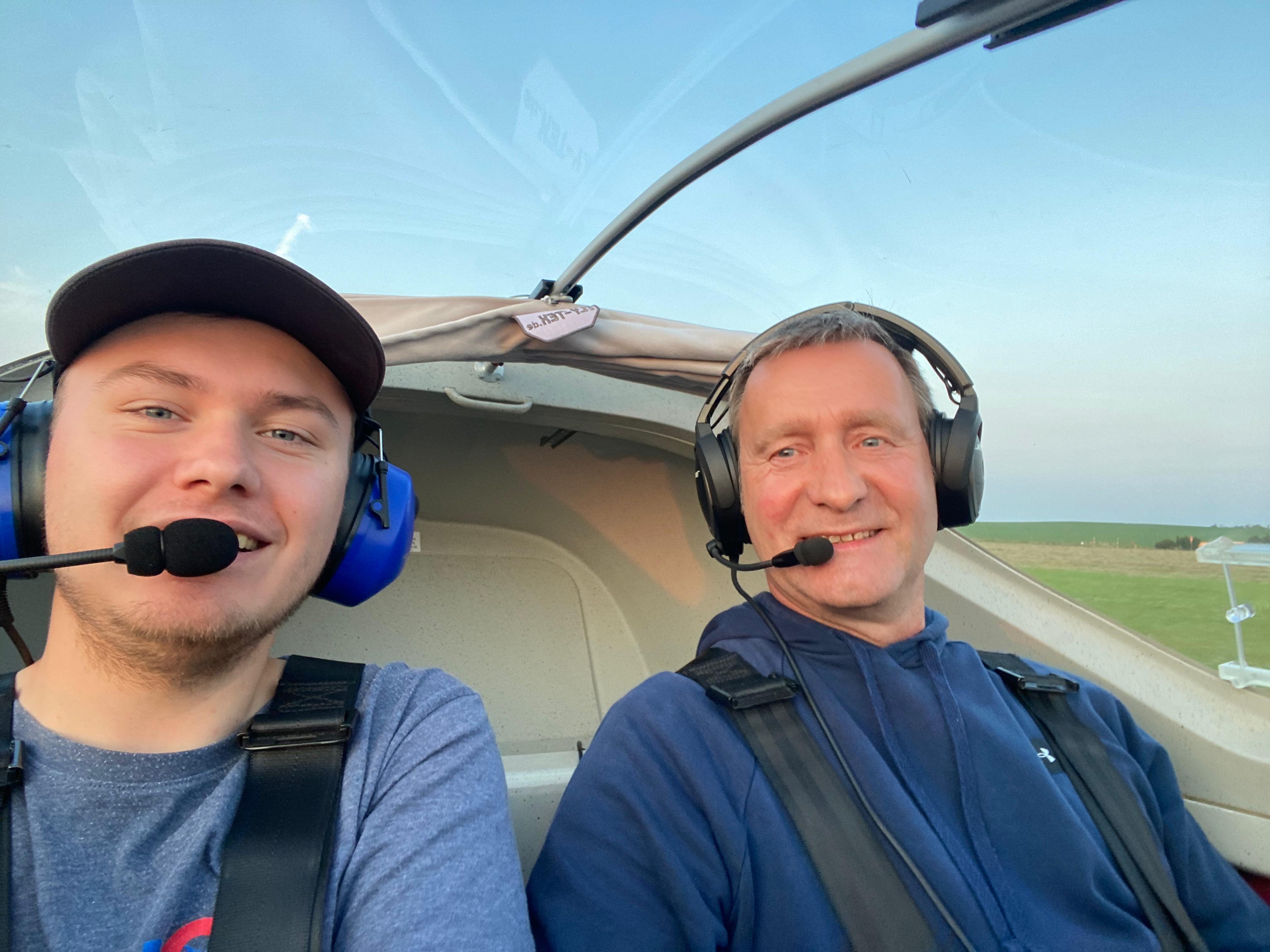 Zwei Männer mit Headsets sitzen in einem Flugzeugcockpit und lächeln in die Kamera