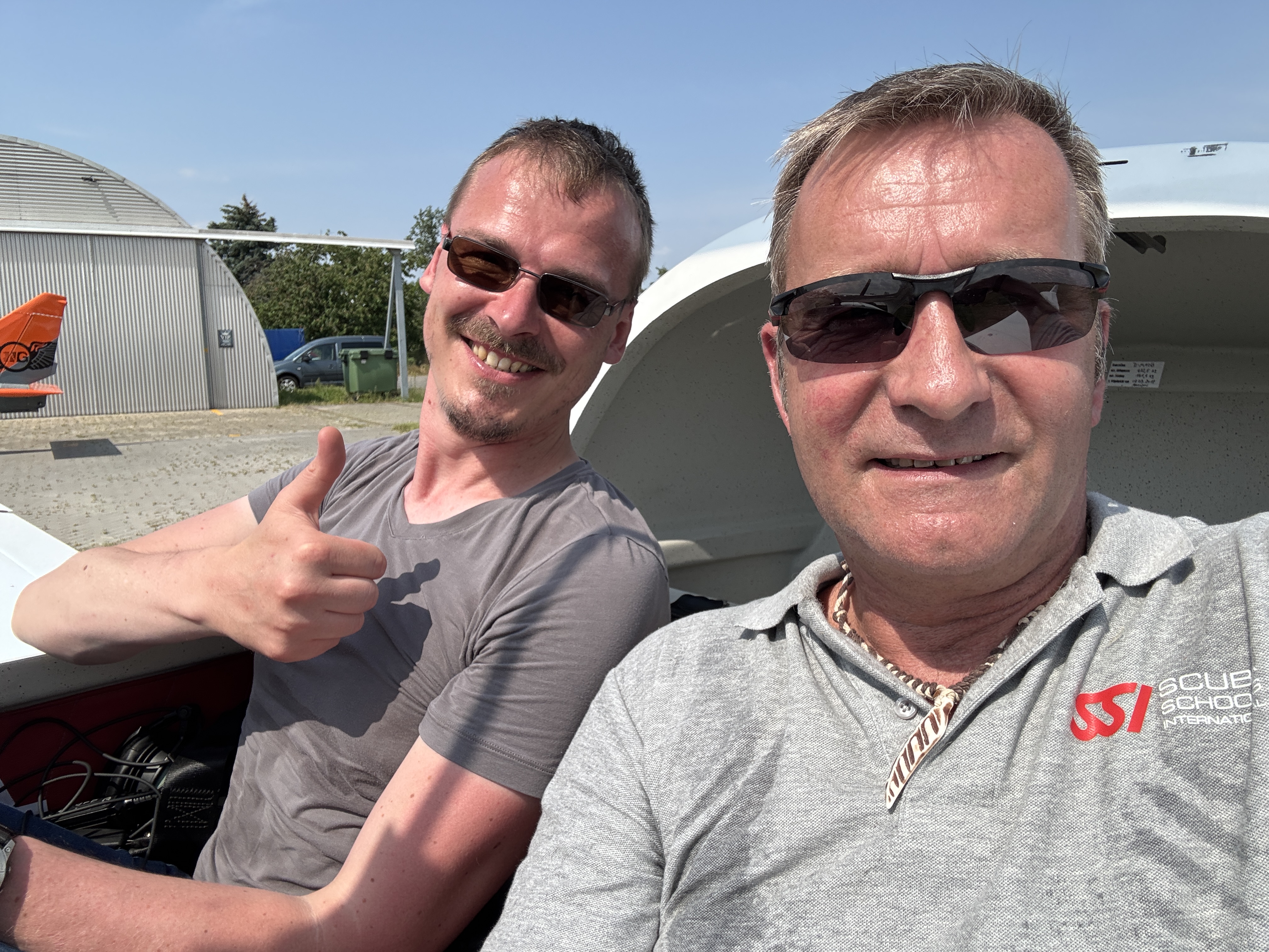 Zwei lächelnde Männer in einem Flugzeug, einer mit Daumen hoch und beide tragen Sonnenbrillen