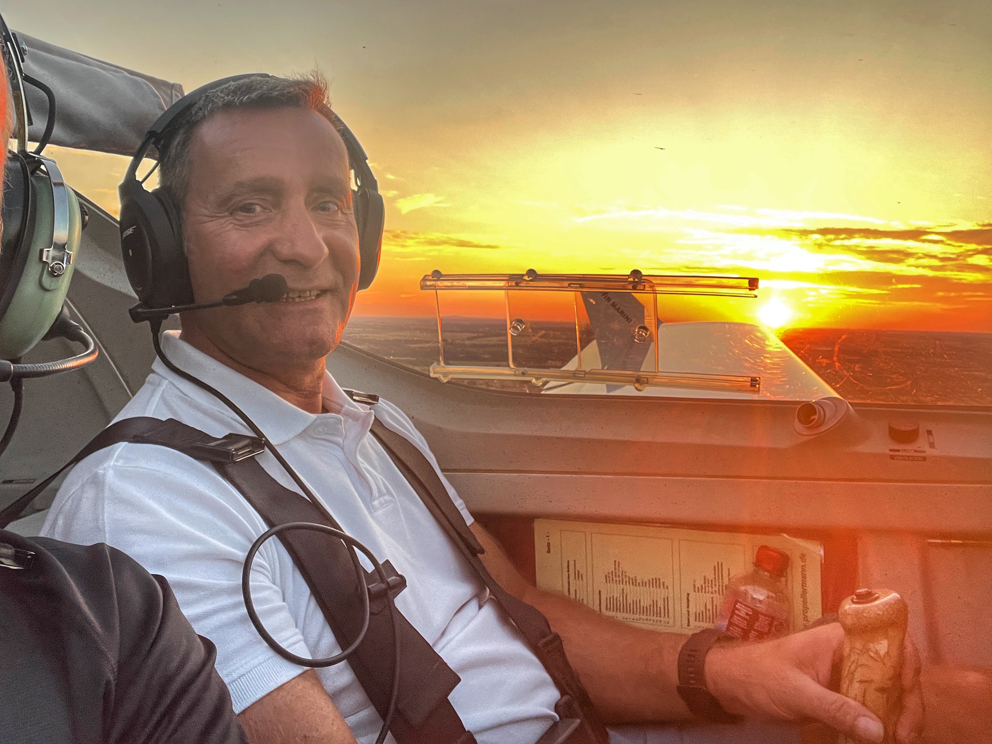 Ein Pilot mit Headset sitzt in einem Flugzeugcockpit und schaut in die Kamera, während draußen die Sonne untergeht.