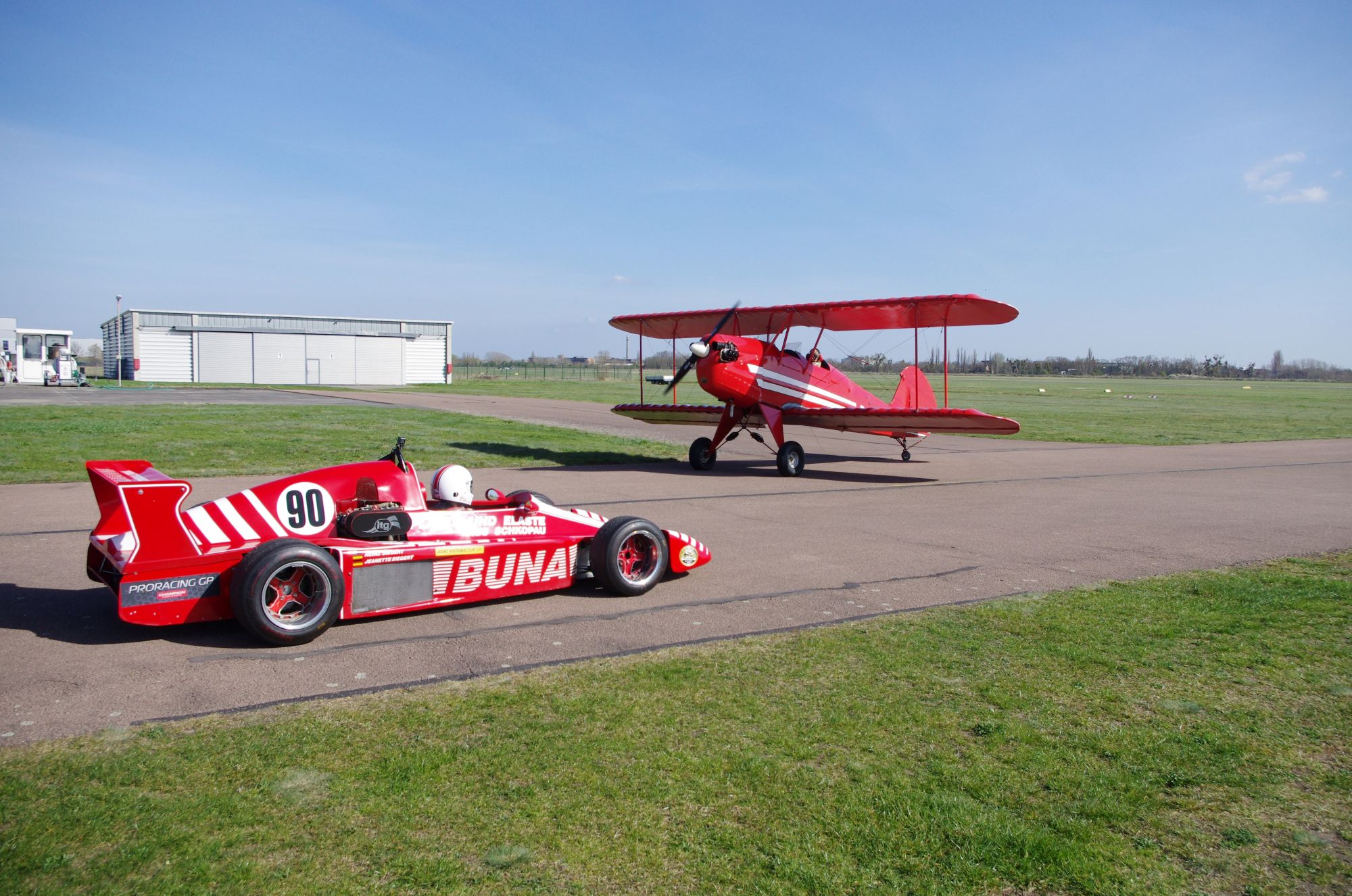 Ein roter Rennwagen neben einem roten Doppeldeckerflugzeug auf einem Flugplatz bei klarem Himmel.