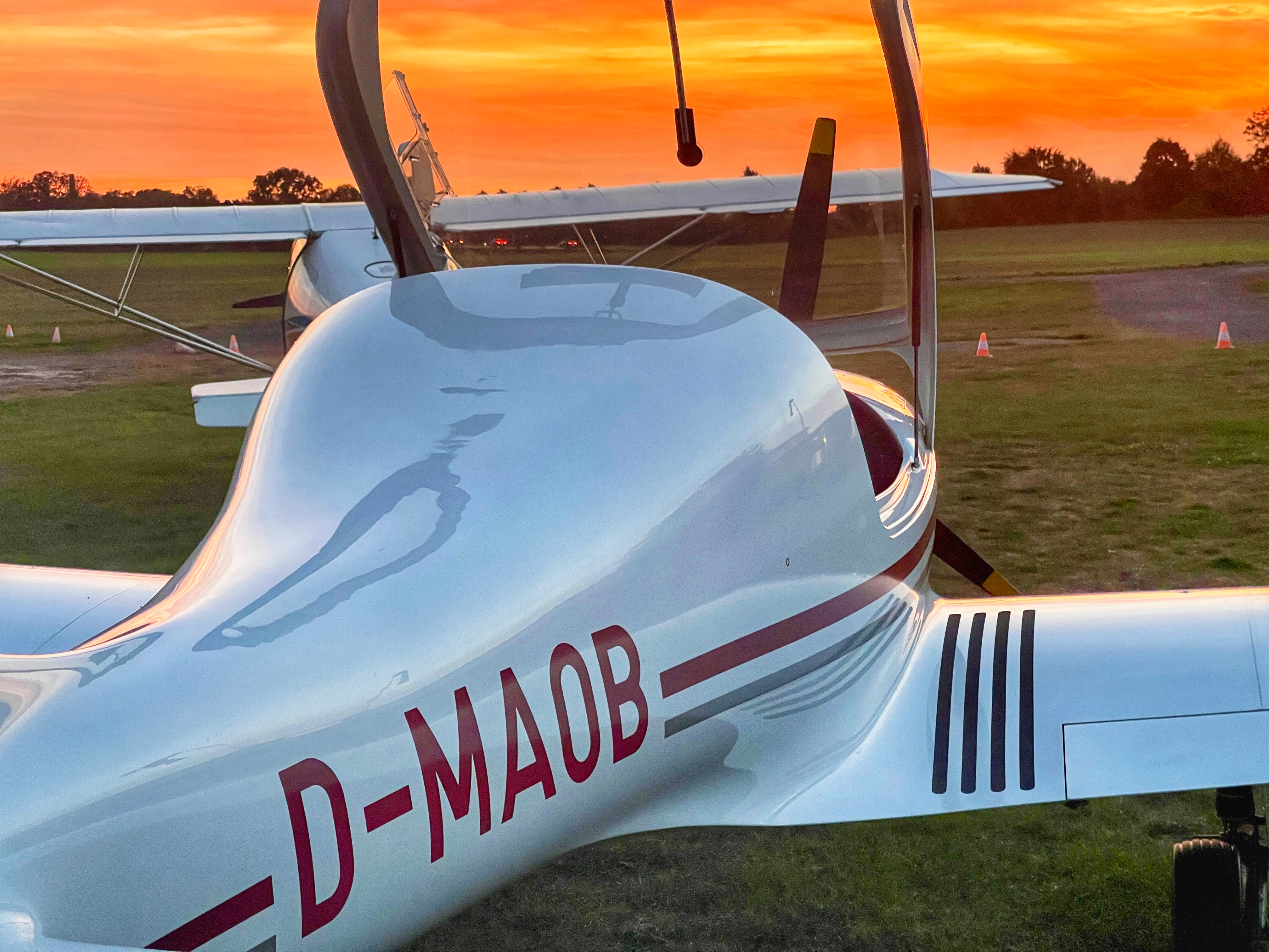 Ein weißes, einmotoriges Leichtflugzeug mit der Kennung D-MAOB steht auf einem Rasenflugplatz, während die Sonne untergeht.