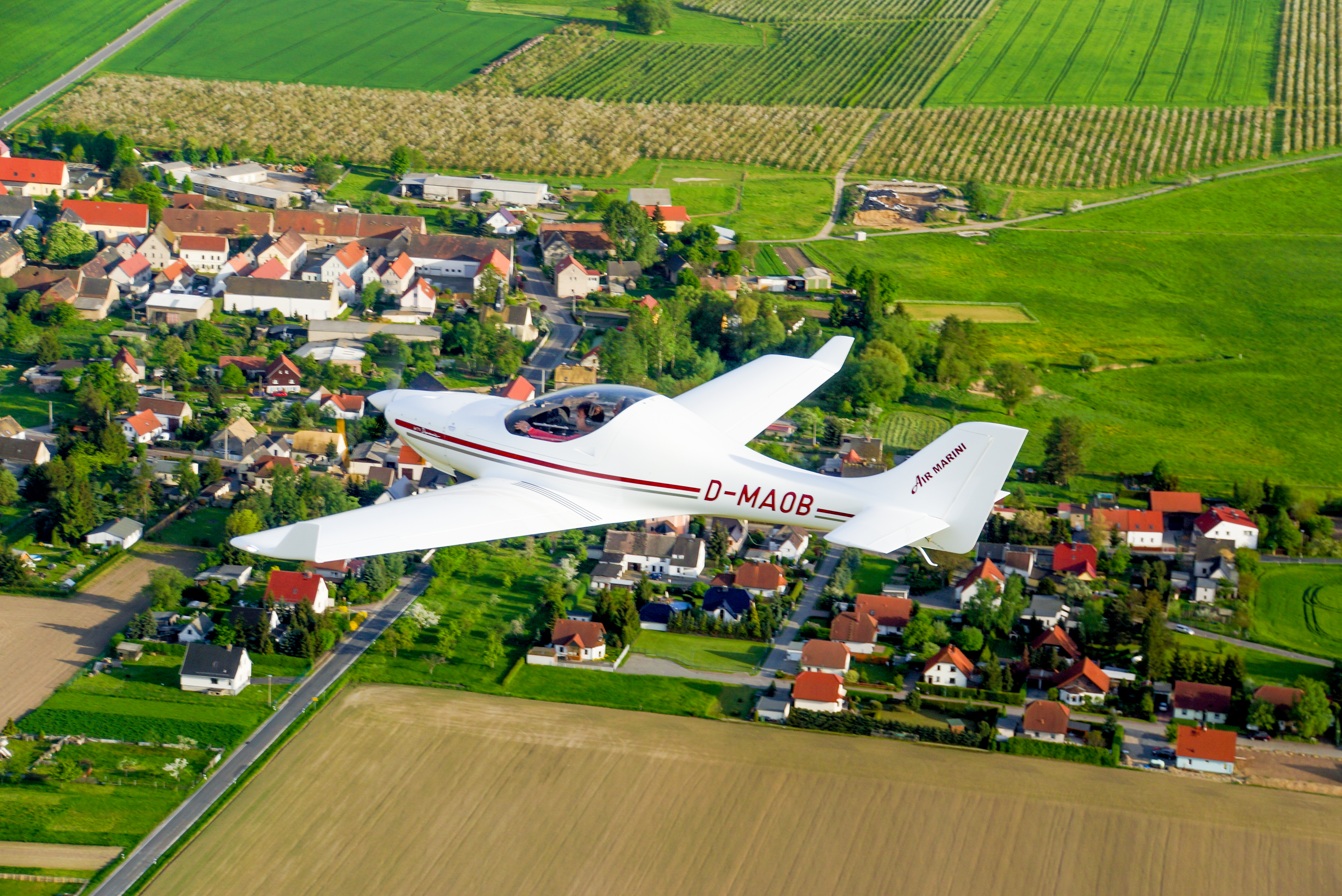 Ein weißes Leichtflugzeug mit der Aufschrift D-MAOB fliegt über einem Dorf und grünen Feldern