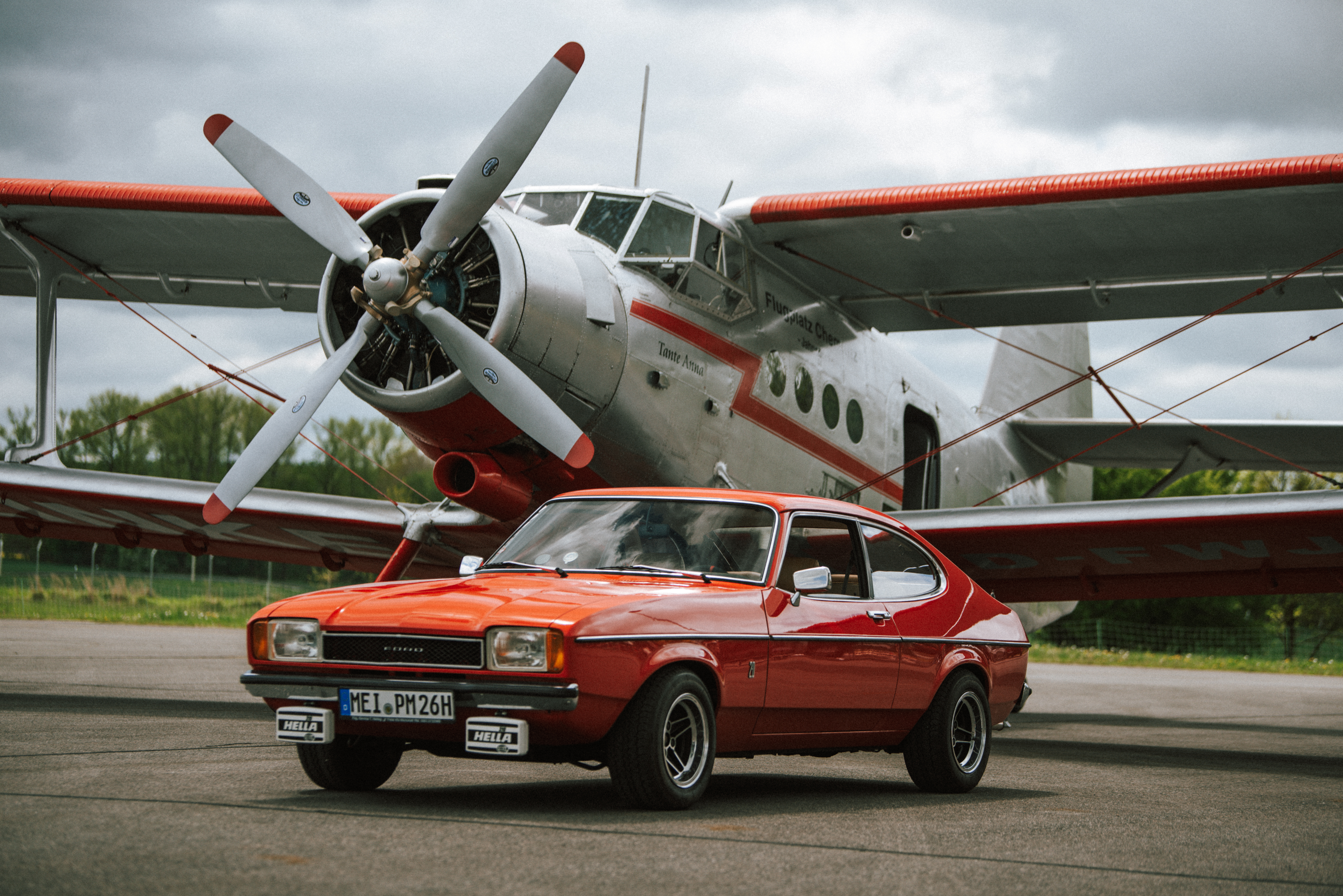 Ein roter Oldtimer-Sportwagen steht stilvoll vor einem großen, weißen Doppeldeckerflugzeug mit roten Akzenten auf einer Landebahn. Der Himmel ist bewölkt, was dem Bild eine nostalgische Atmosphäre verleiht.