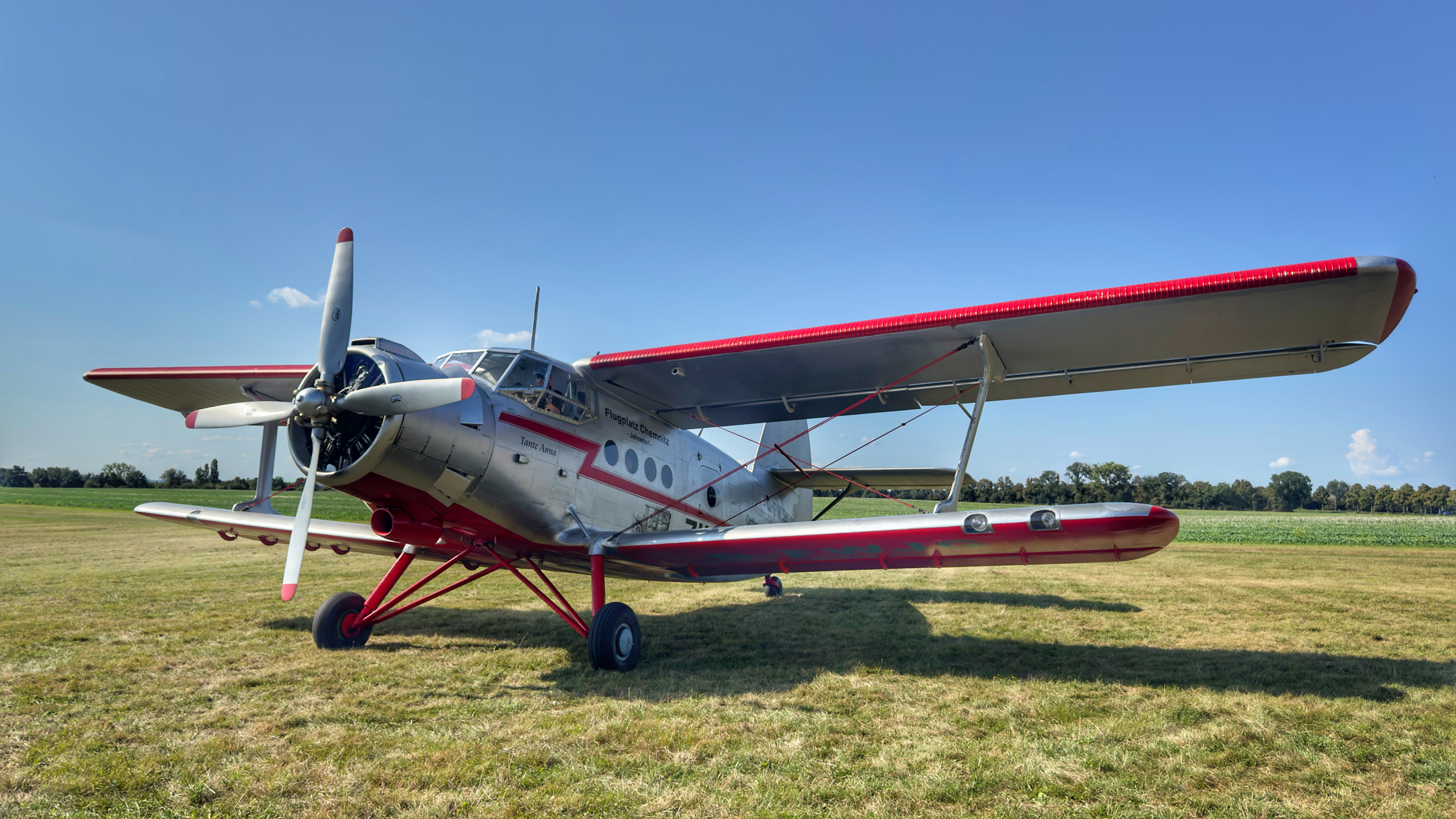 Ein silberrotes Doppeldecker-Flugzeug steht auf einer Wiese. Der Himmel ist klar und blau.