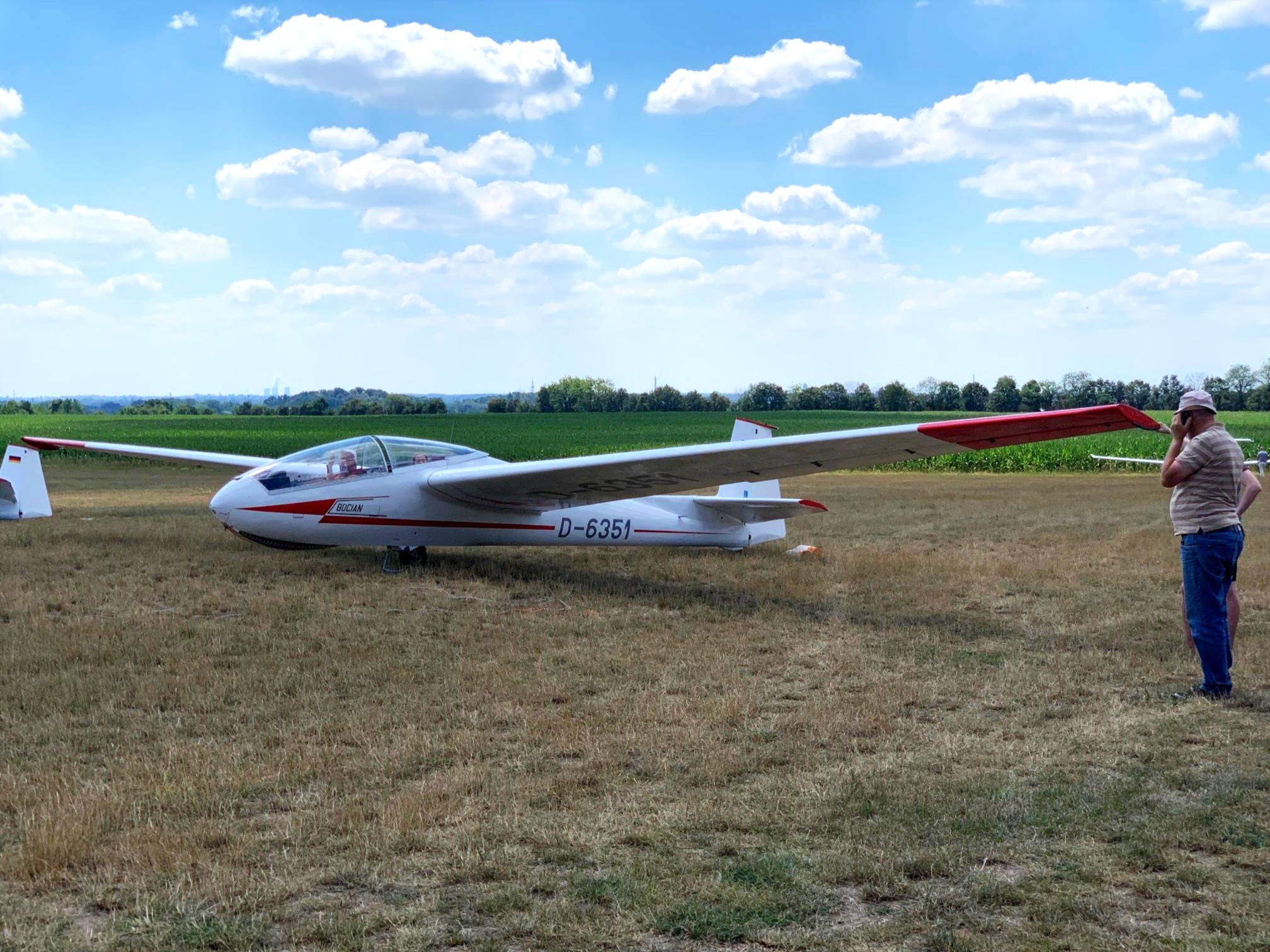 Ein weißes Segelflugzeug mit roter Markierung steht auf einem trockenen Feld. Ein Mann steht daneben und schaut in die Richtung des Flugzeugs. Im Hintergrund sind Felder und ein blauer Himmel mit einigen Wolken zu sehen.