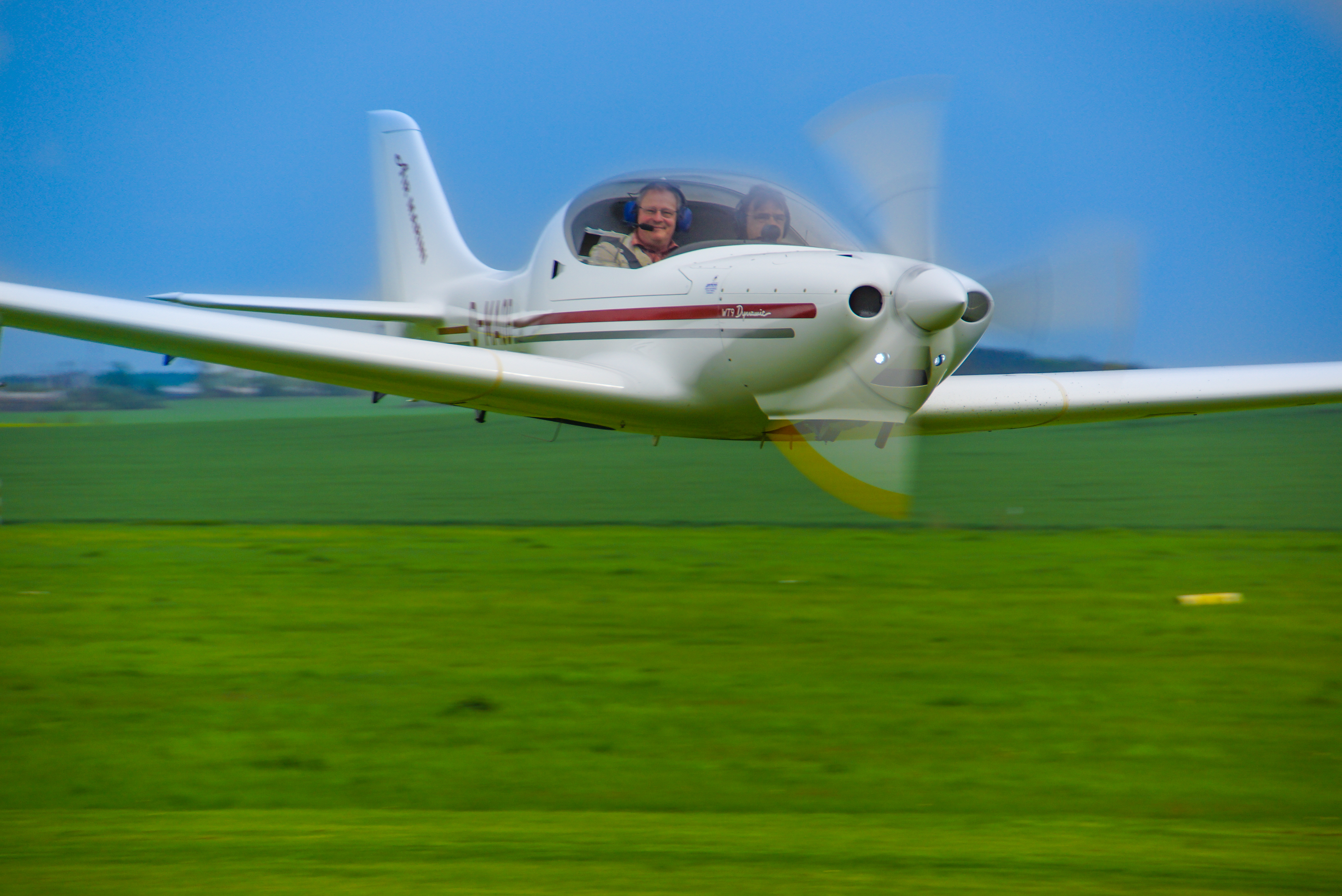 Ein weißes, zweisitziges Leichtflugzeug mit drehendem Propeller fliegt über ein grünes Feld, zwei Personen im Cockpit