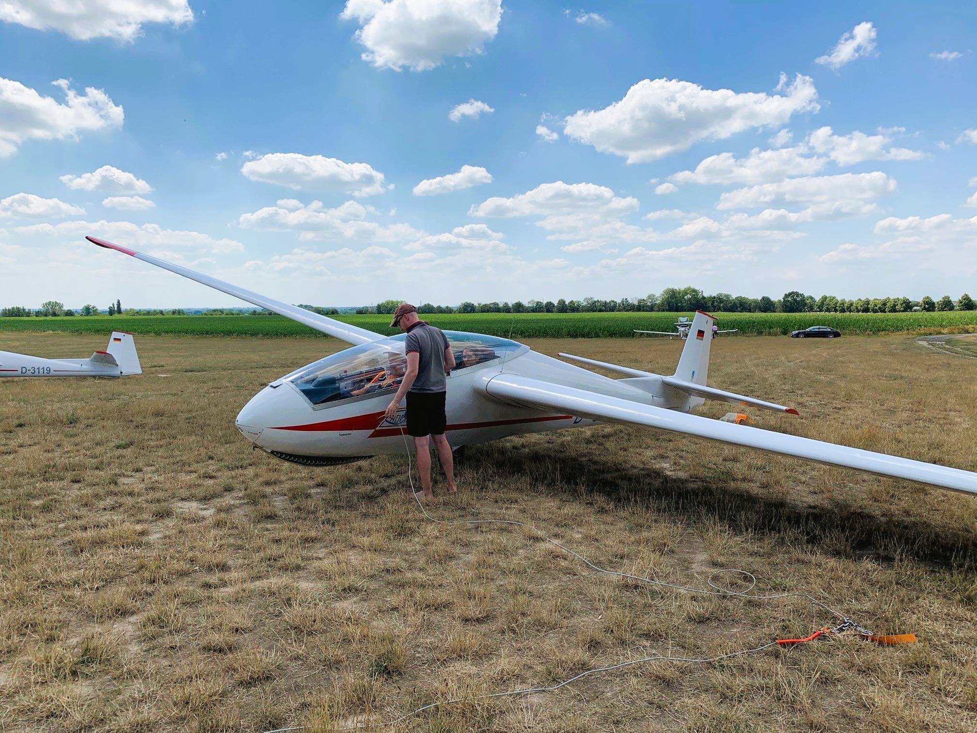 Person überprüft ein Segelflugzeug auf einer Wiese unter einem blauen Himmel mit weißen Wolken