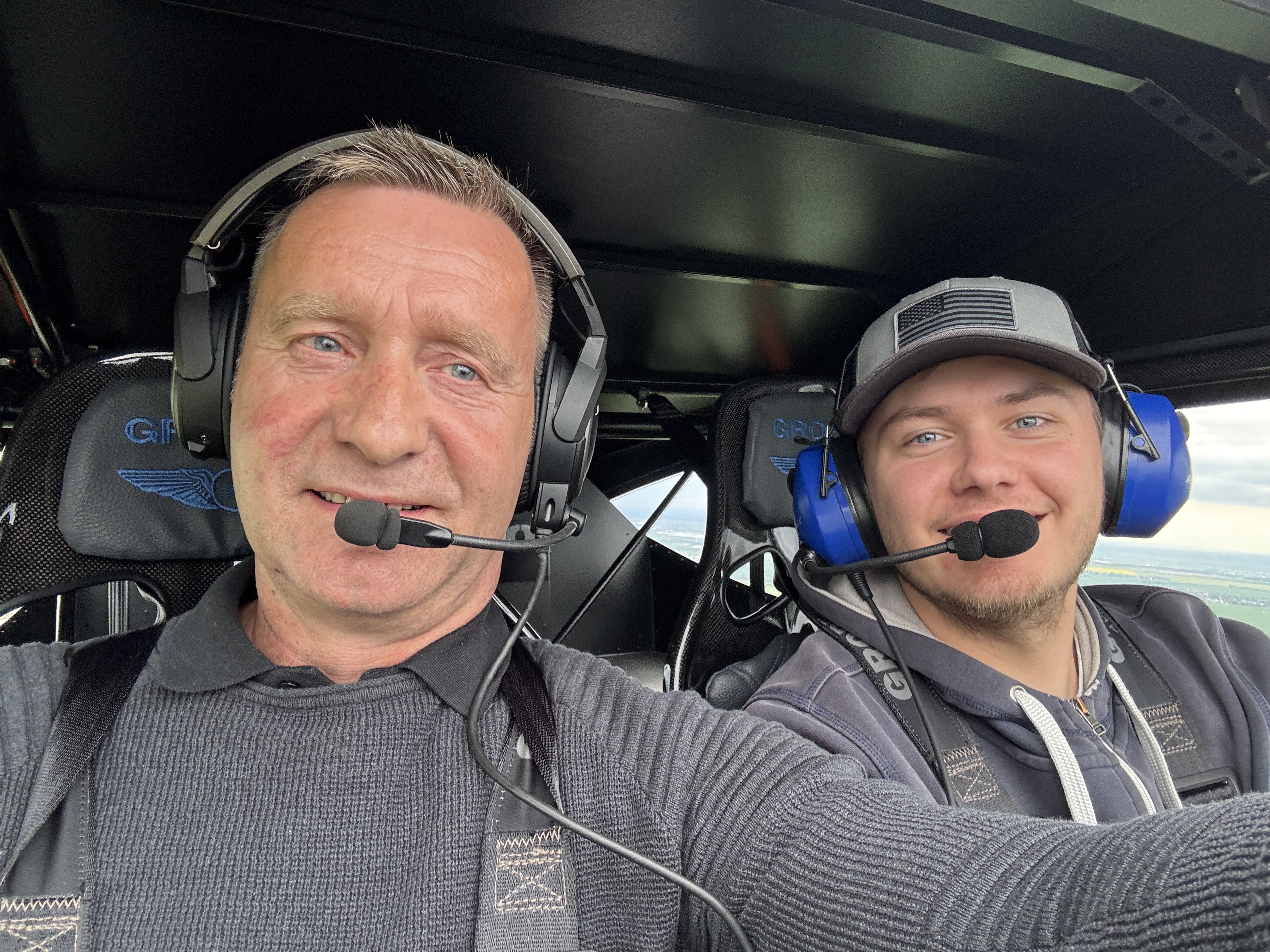 Zwei Männer mit Headsets sitzen im Cockpit eines Flugzeugs und lächeln in die Kamera.