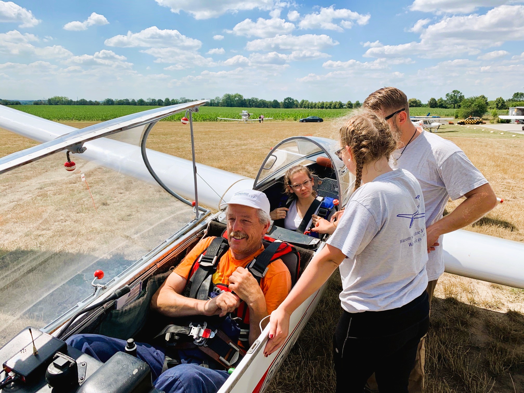 Menschen helfen einem Piloten, sich in einem Segelflugzeug vorzubereiten, bevor es vom Grasfeld aus startet, bei sonnigem Wetter mit blauem Himmel.