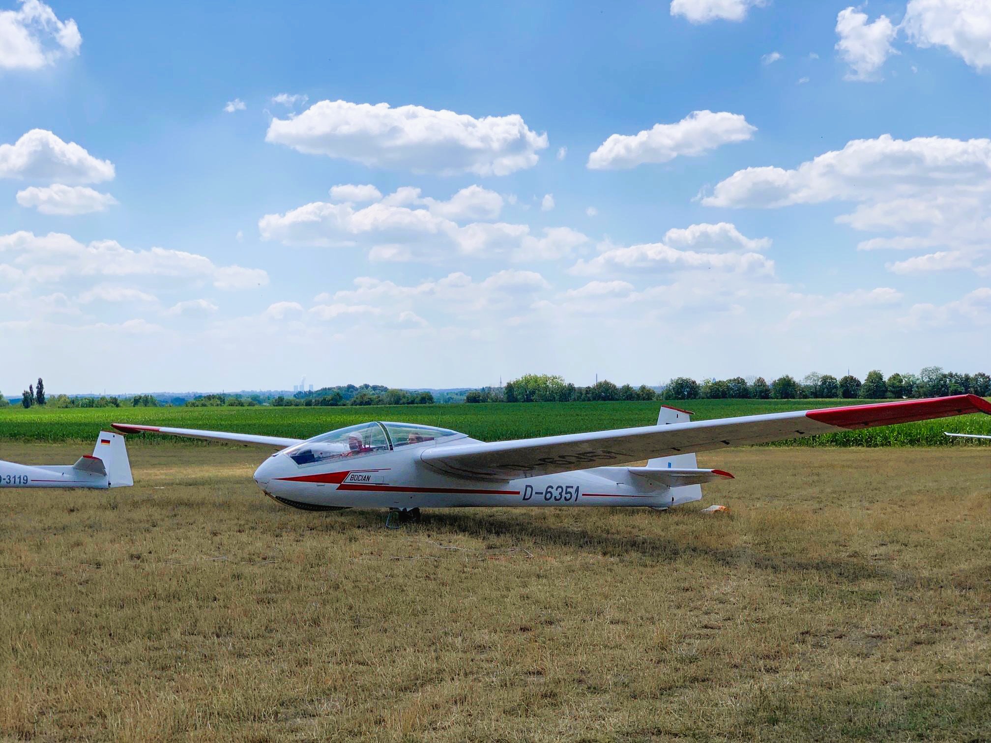 Ein weiß-rotes Segelflugzeug steht auf einer grasbewachsenen Fläche vor einem blauen Himmel mit einigen Wolken.