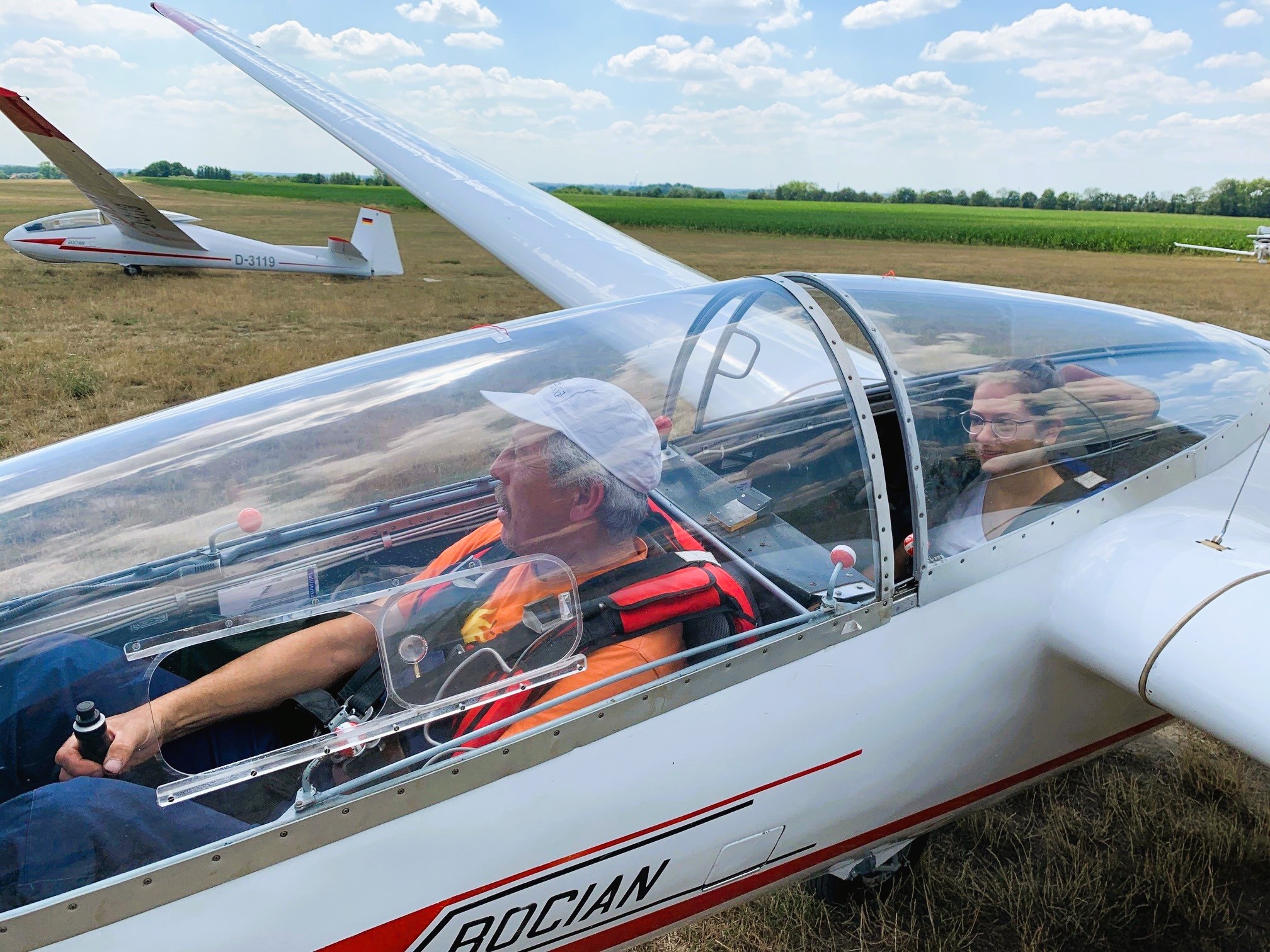 Zwei Personen sitzen in einem Segelflugzeug auf einem Feld, ein weiteres Segelflugzeug ist im Hintergrund sichtbar.
