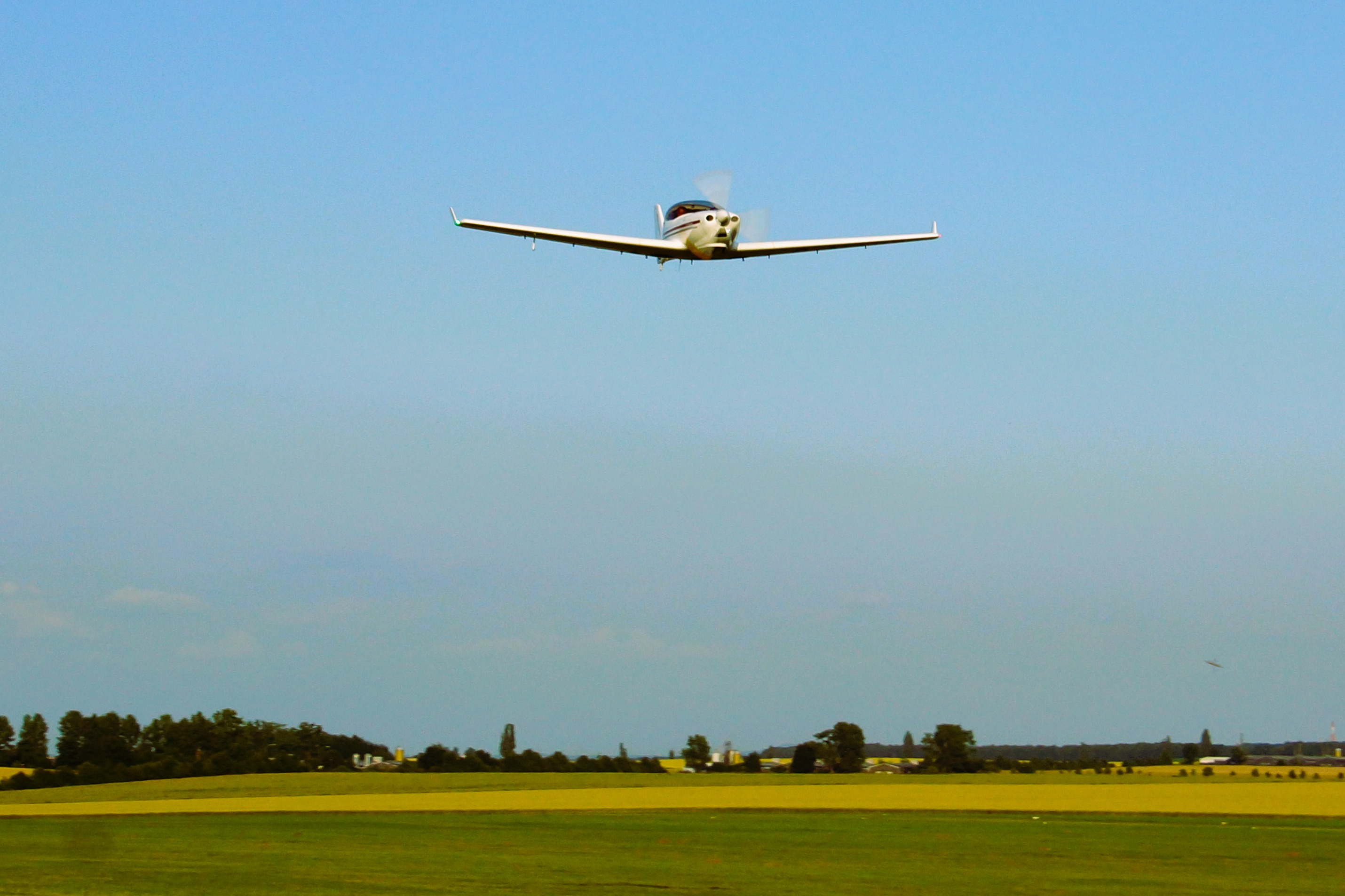 Ein Kleinflugzeug im Anflug auf eine Landebahn, über eine grüne Wiese fliegend, mit blauem Himmel im Hintergrund.