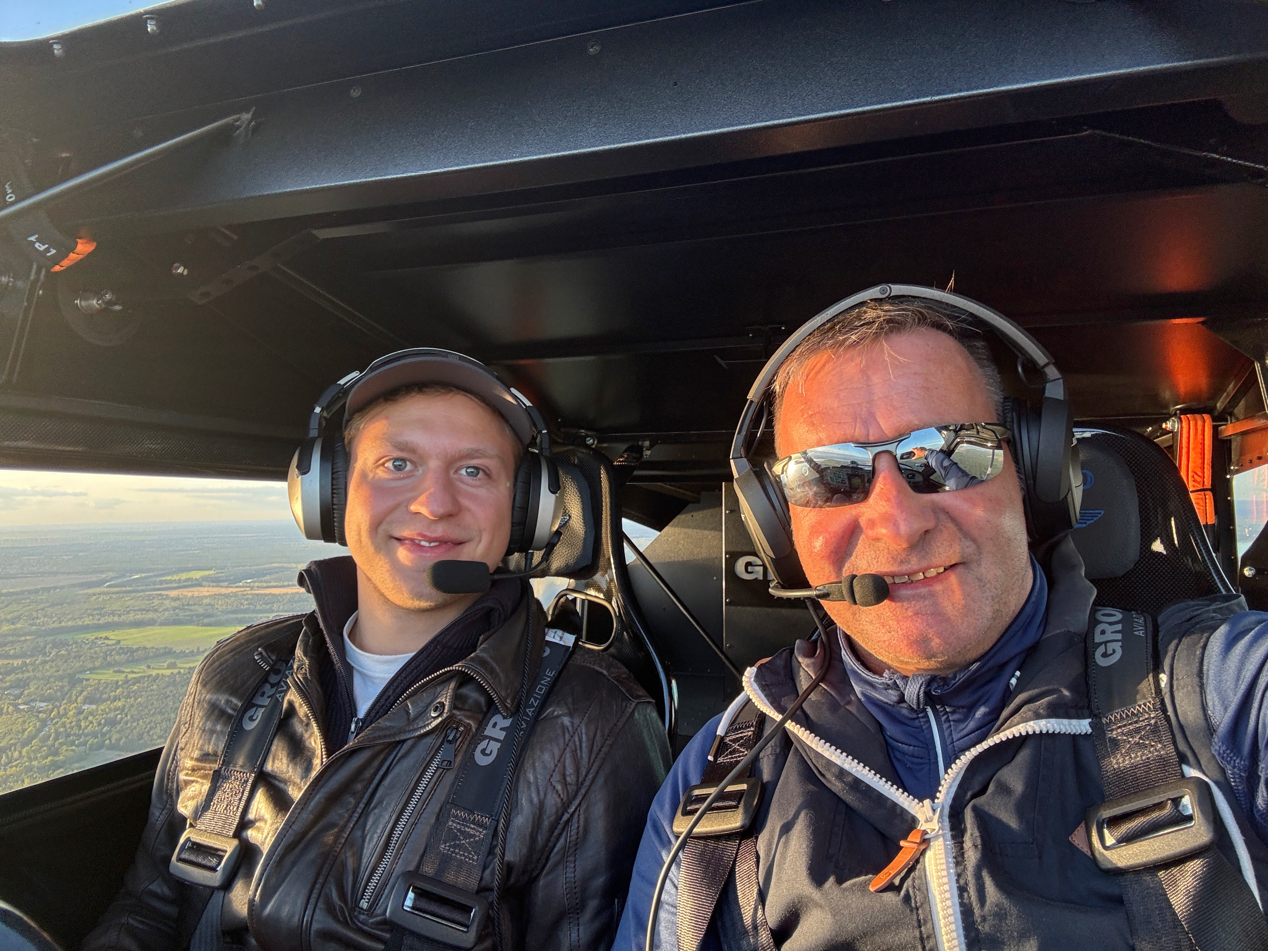 Zwei Männer mit Headsets im Cockpit eines kleinen Flugzeugs, mit Sicht auf die Landschaft durch das Fenster