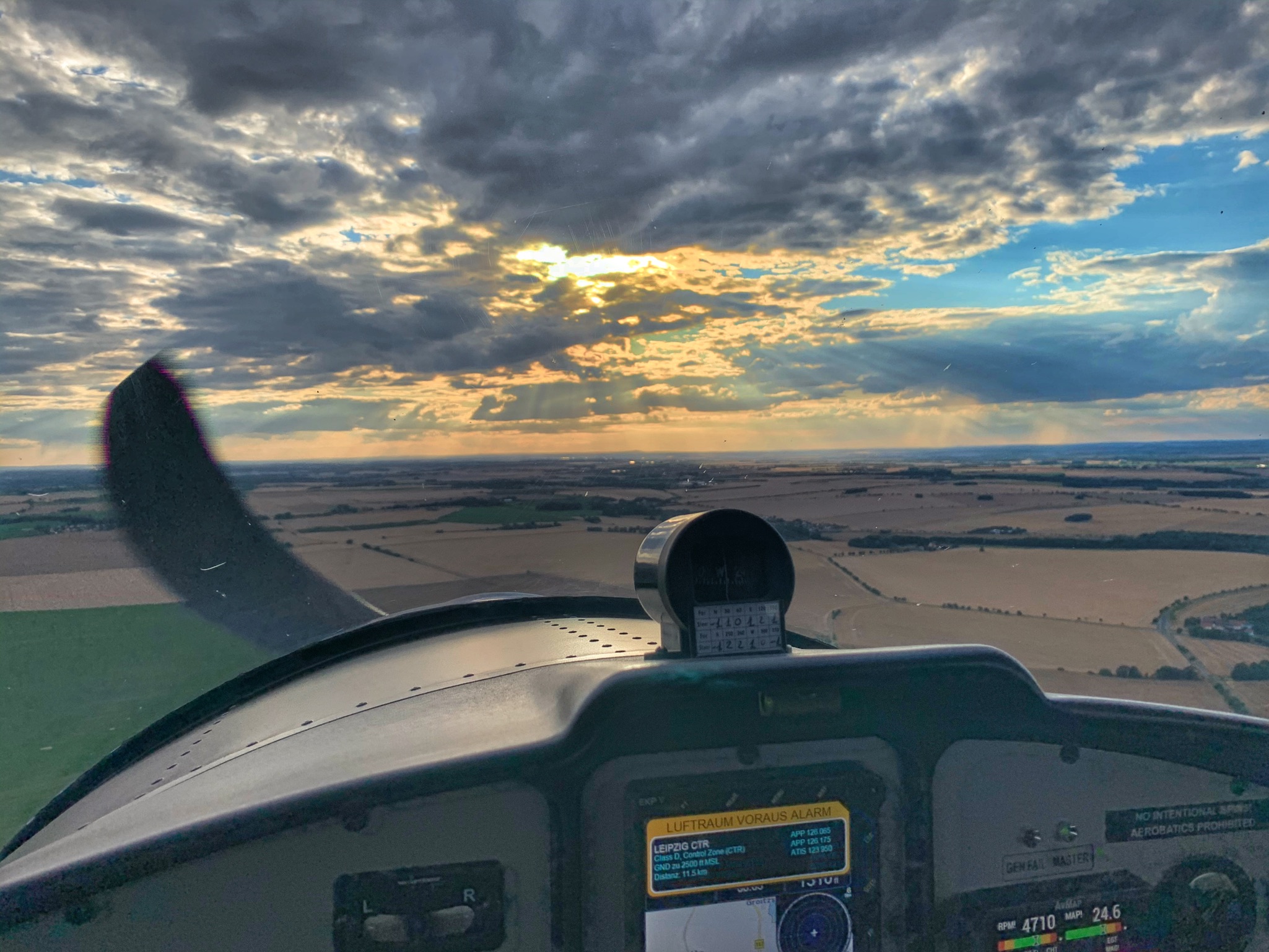 Blick aus dem Cockpit eines Kleinflugzeugs auf eine Landschaft bei Sonnenuntergang mit dramatischen Wolken.