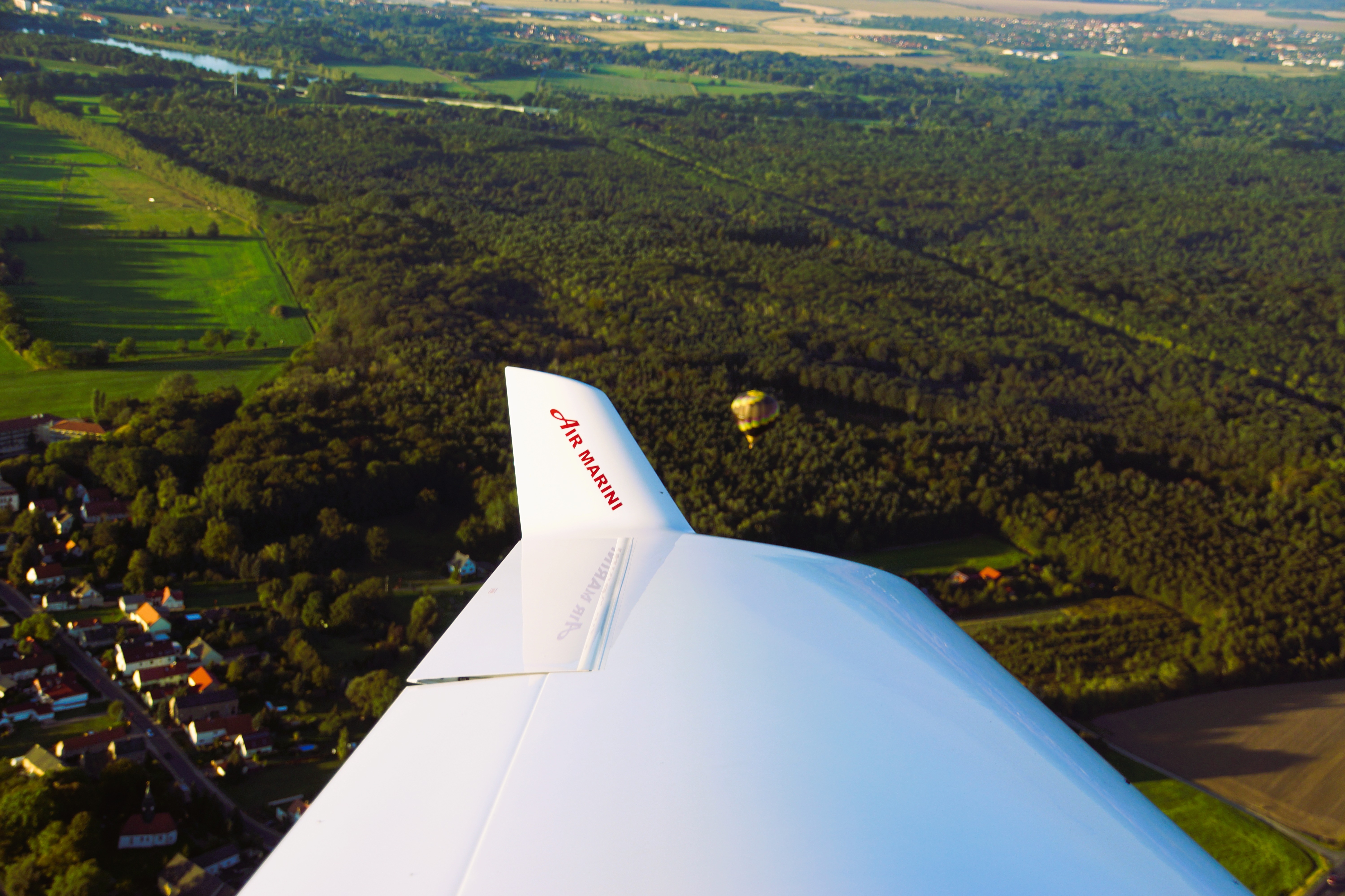 Blick auf den Flugzeugflügel über einer grünen, bewaldeten Landschaft mit einem Dorf im Hintergrund